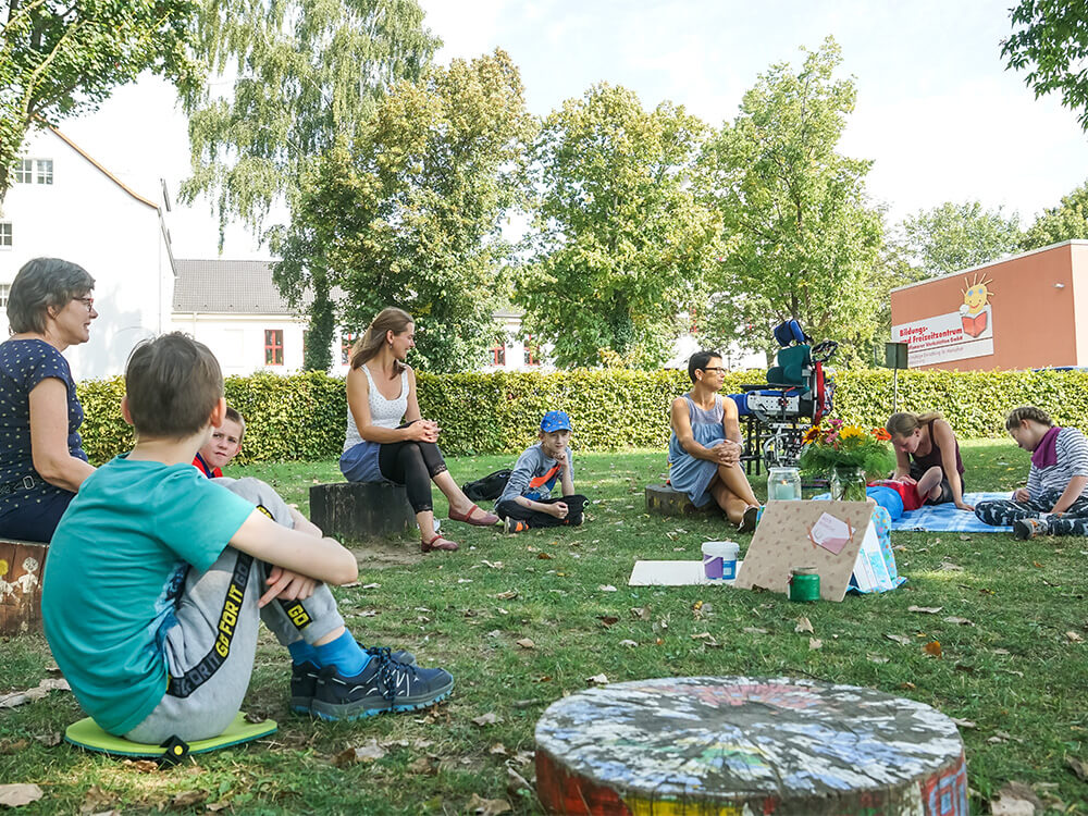 Kinder und Lehrkräfte sitzen in einer Runde auf dem Rasen